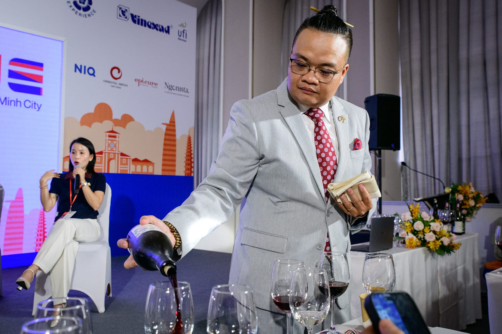 Sommelier pouring red wine for the tasting session at the Next Generation Wine Consumer panel, Wines Experience Ho Chi Minh City 2026, with event sponsor logos including NielsenIQ and UFI visible in the background